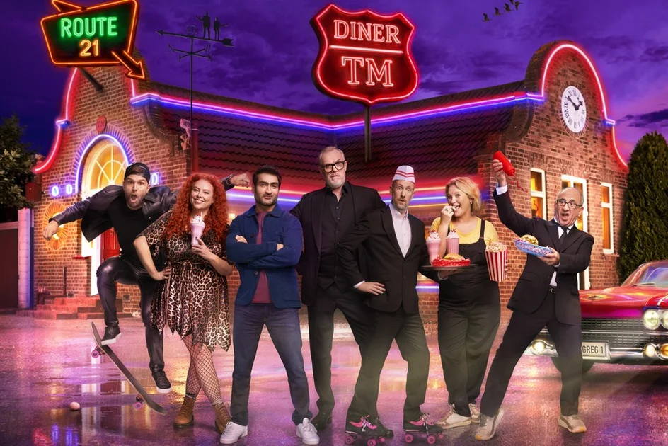 Joel Dommett, Amy Gledhill, Kumail Nanjiani, Greg Davies, Alex Horne, Joanna Page and Armando Iannucci standing in front of the Taskmaster house with an American diner aesthetic. Alex is dressed as a serving waiter on roller skates, while the cast are holding milkshakes, burgers and popcorn.