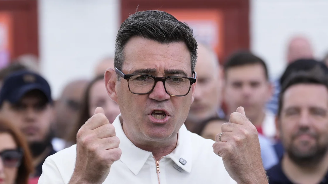 Andy Burnham making a speech at Ashton United FC in Ashton-under-Lyme, Greater Manchester. His hands are clenched and he is mid-speech. He is wearing a white, collared, short sleeved shirt with a zipper. He has glasses.