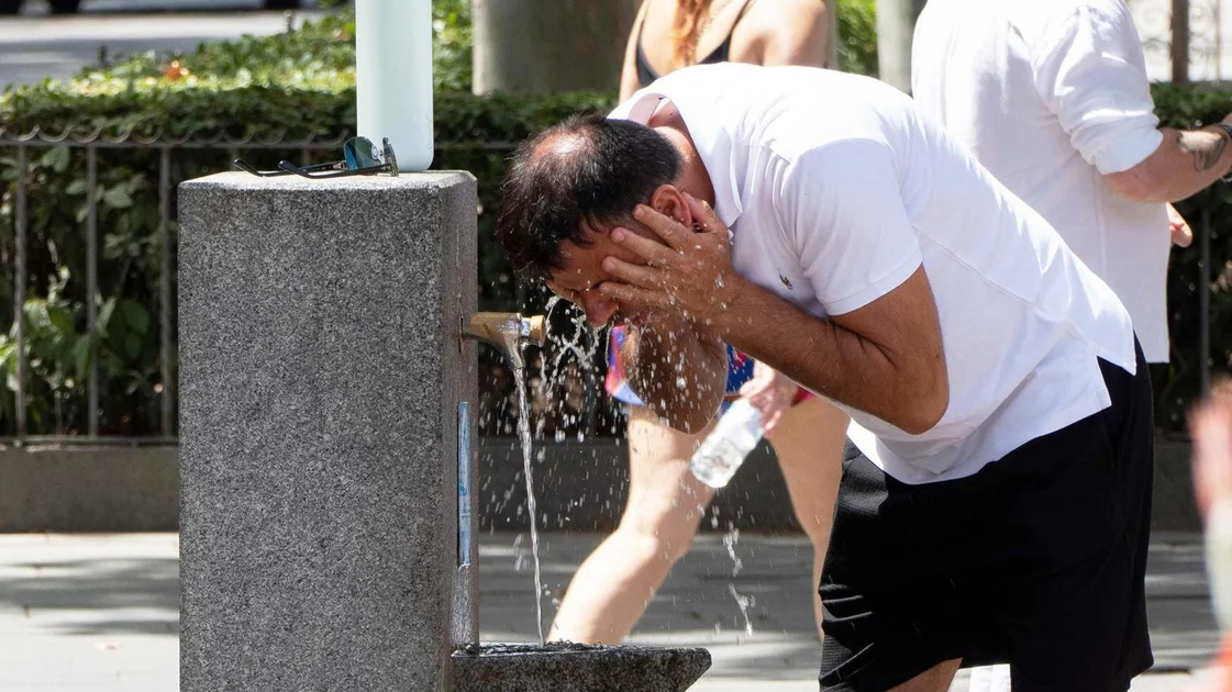 A man splashes his face with water at a drinking fountian in Spain during the heatwave. He is wearing a white T-shirt
