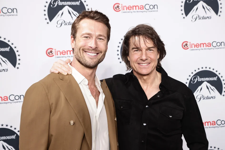Glen Powell in a tan blazer and Tom Cruise in a black shirt smiling together in front of a Paramount Pictures and CinemaCon step-and-repeat backdrop.
