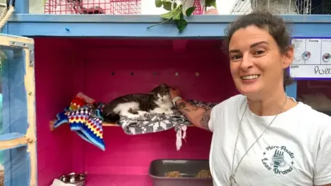 Woman standing in front of a cat shed, holding a cat with her right hand. The white and pepper black cat is on a rug.