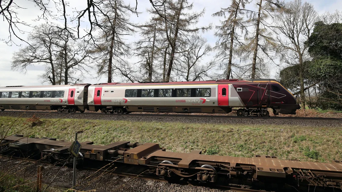 The cab and first carriage of a CrossCountry train travels on tracks in the countryside. In the background trees and fields can be seen.